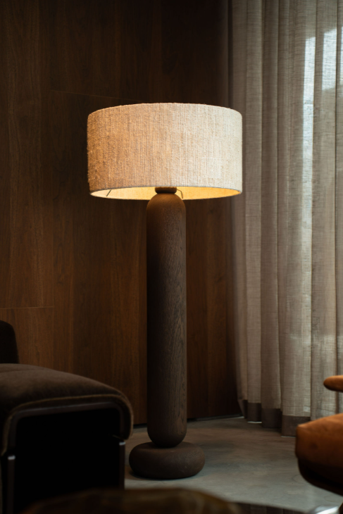 Vloerlamp met donkere houten voet en naturel geweven kap tegen een notenhouten wand, met linnen gordijnen ernaast.