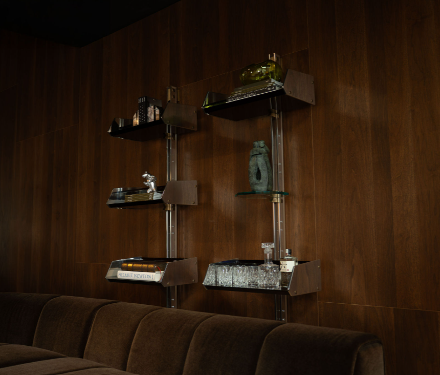 Metalen wandplanken met boeken, kristallen glazen, karaf en sculptuur aan een notenhouten wand boven een bruine fluwelen bank.