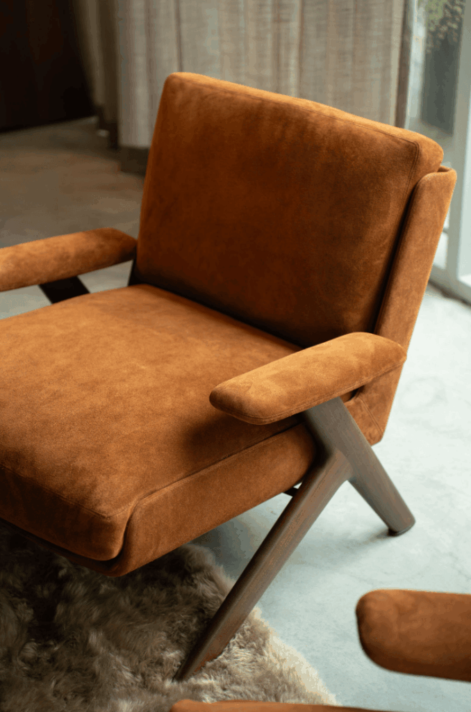 Luxe fauteuil in warme cognackleurige stoffering met houten frame en armleuningen, gecombineerd met een zacht vloerkleed bij Noort Interieur in Noordwijk.
